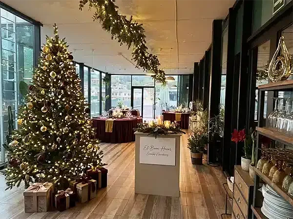 Salón de eventos con amplias cristaleras, decorado con un árbol de Navidad y luces cálidas, preparado para comidas de empresa.